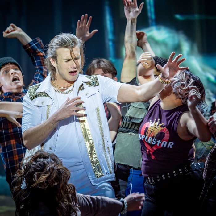 A cast of students performs in a College of the Arts production of Jesus Christ, Superstar with the spotlight on the actor playing Jesus Christ singing to his followers who surround him