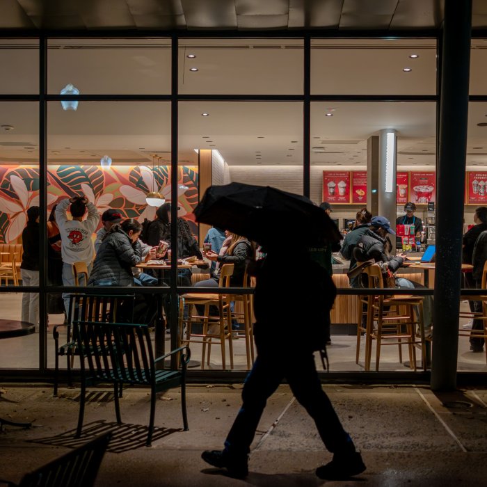 The silhouette of a student at night with an umbrella passes in front of a lit-up wall of windows at Cafe Diem, a campus coffee house bustling with activity