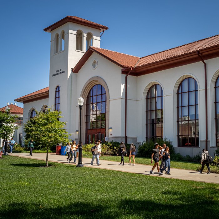 Students walk in front of Montclair State University’s School of Communication and Media on a sunny day, with large arched windows and red-tiled roof visible in the background.