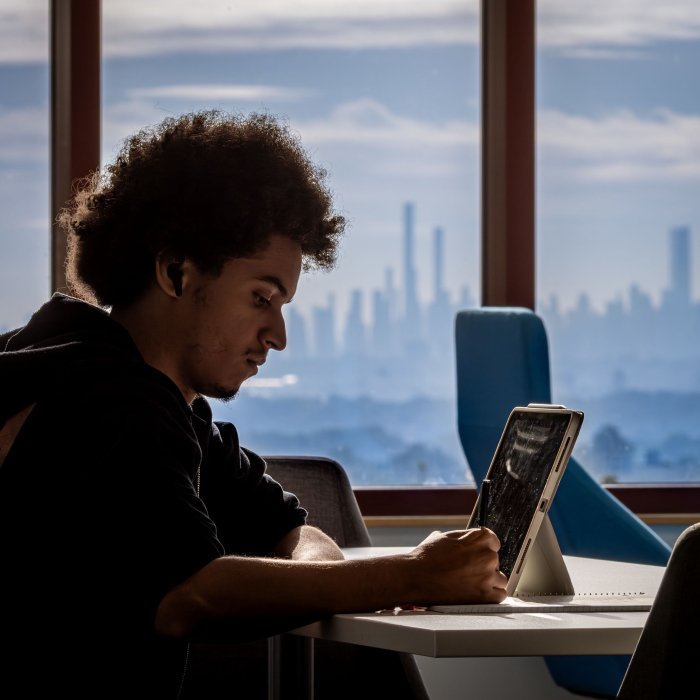 Student studies seated by a window with the NYC skyline in background.