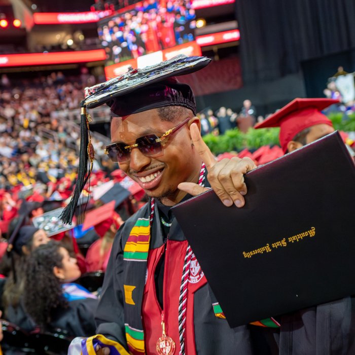 a graduate in black cap, gown and sunglasses smiles while holding diploma holder