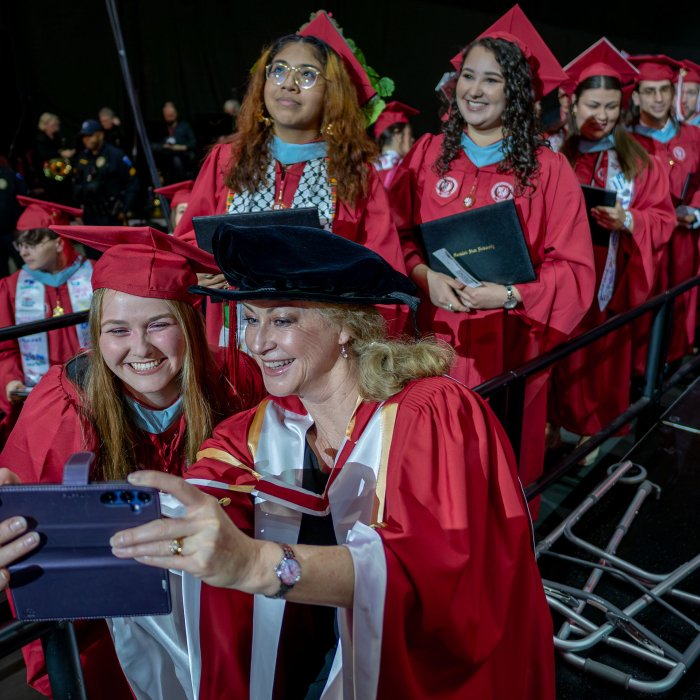 A faculty member in red gown poses for a selfie with a graduate.