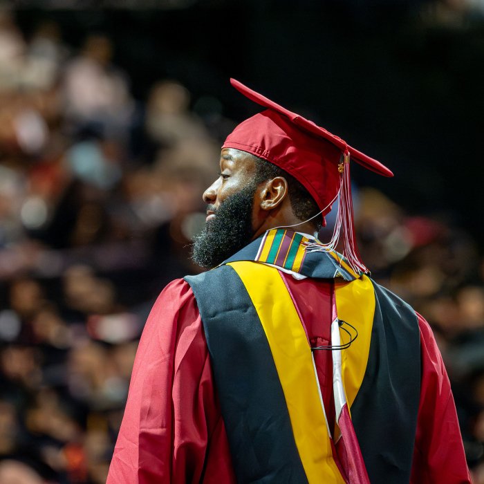 A master's graduate in red cap and gown looks toward the crowd from the stage