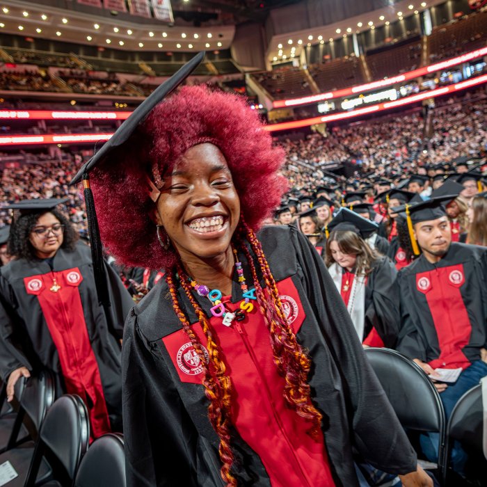 A smiling undergraduate in a black gown and cap walks between rows of seated classmates, her bright red hair and colorful beaded necklace standing out.