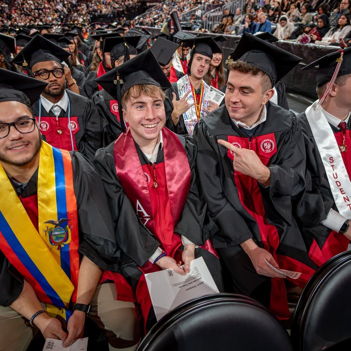 Four undergraduate students in black gowns and caps sit together in a row, smiling and laughing, with one student playfully pointing at a classmate.