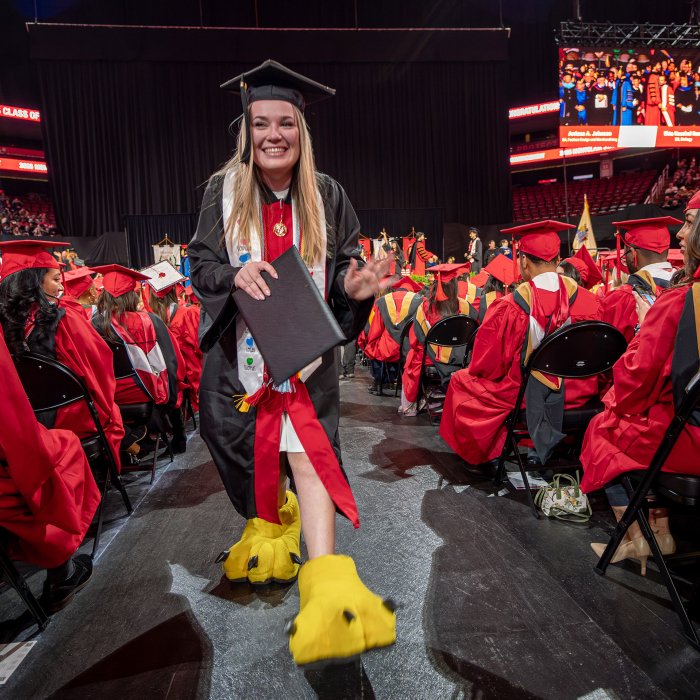 An undergraduate in a black gown and cap walks down the aisle holding her diploma cover while wearing oversized yellow mascot feet, continuing the tradition in which students who served as the college mascot reveal themselves at graduation.