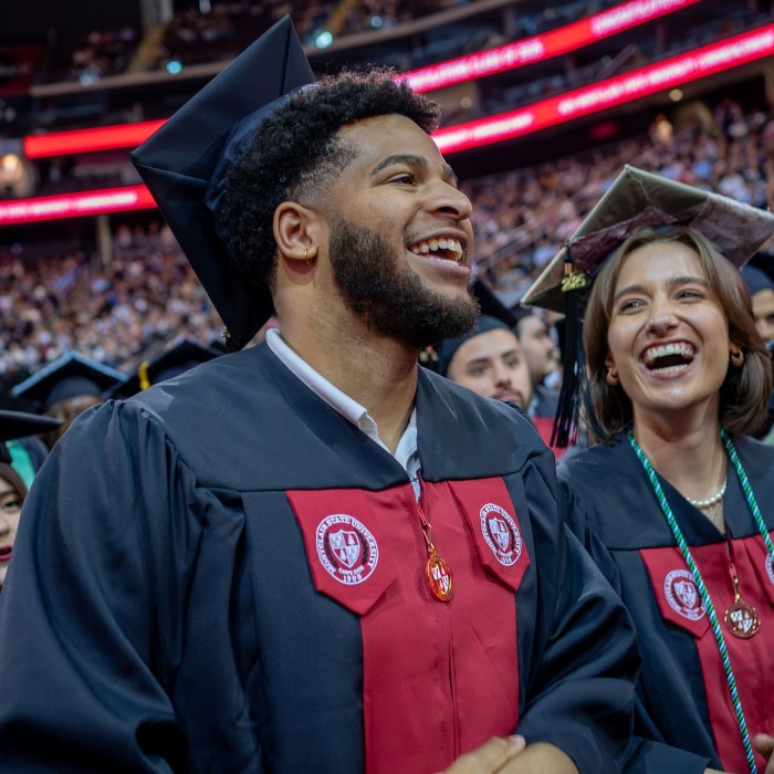 Two graduates in black caps and gowns, smiling in the crowd of fellow graduates.