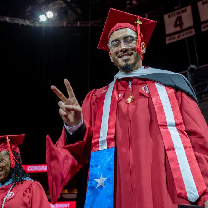 master's graduate in red cap and gown smiling and making a peace sign