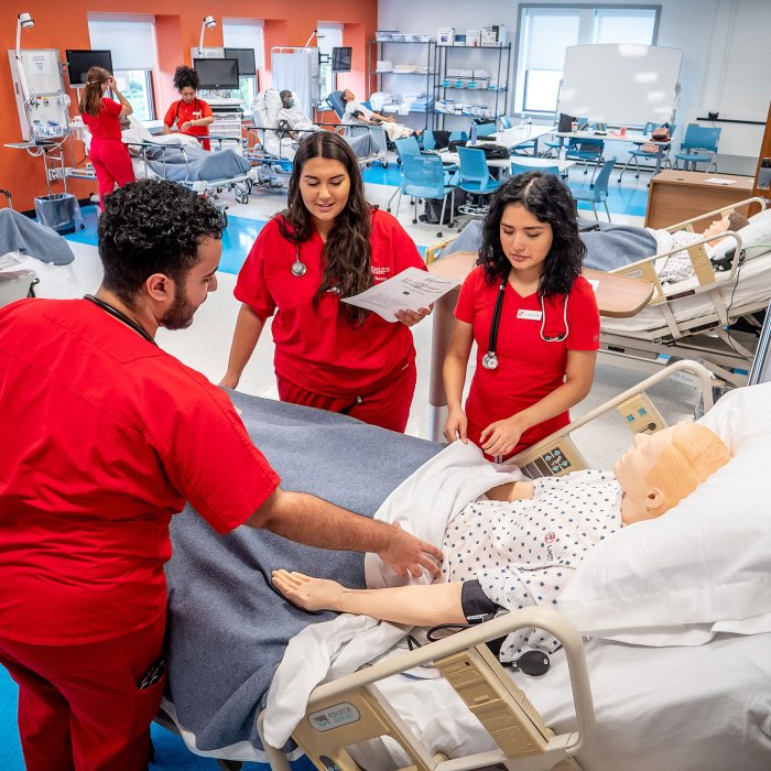 Three nursing students taking notes over a medical mannequin in hospital bed