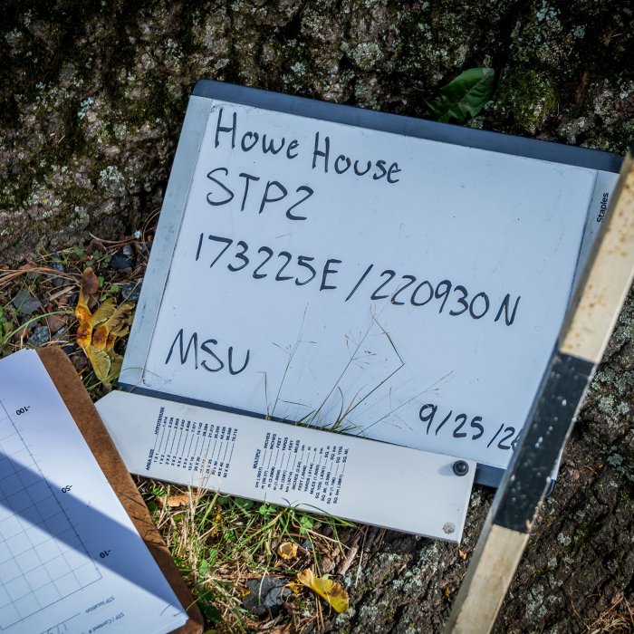A whiteboard sign, a clipboard and a measuring stick lean against a tree.