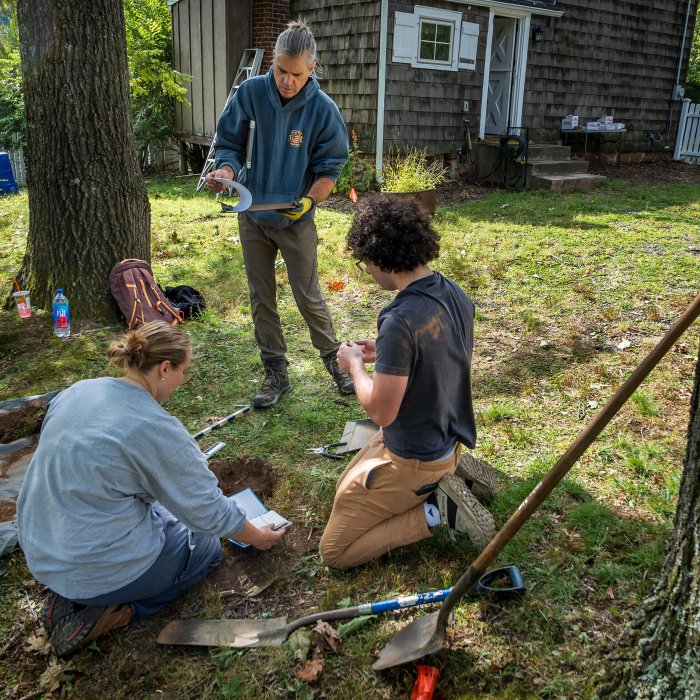 Three people stand or kneel by a hole in the ground with digging tools nearby.