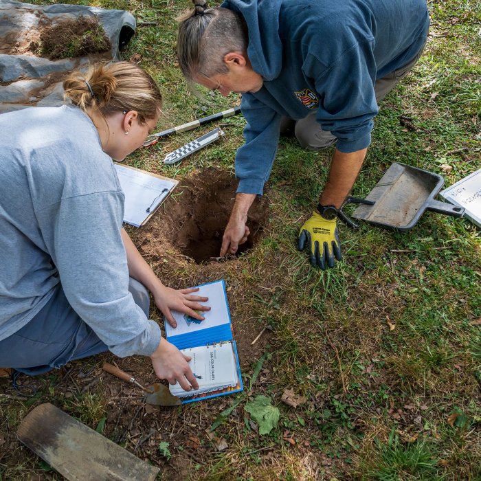 A woman with a notebook kneels beside a hole in the ground while a kneeling man points into the hole.