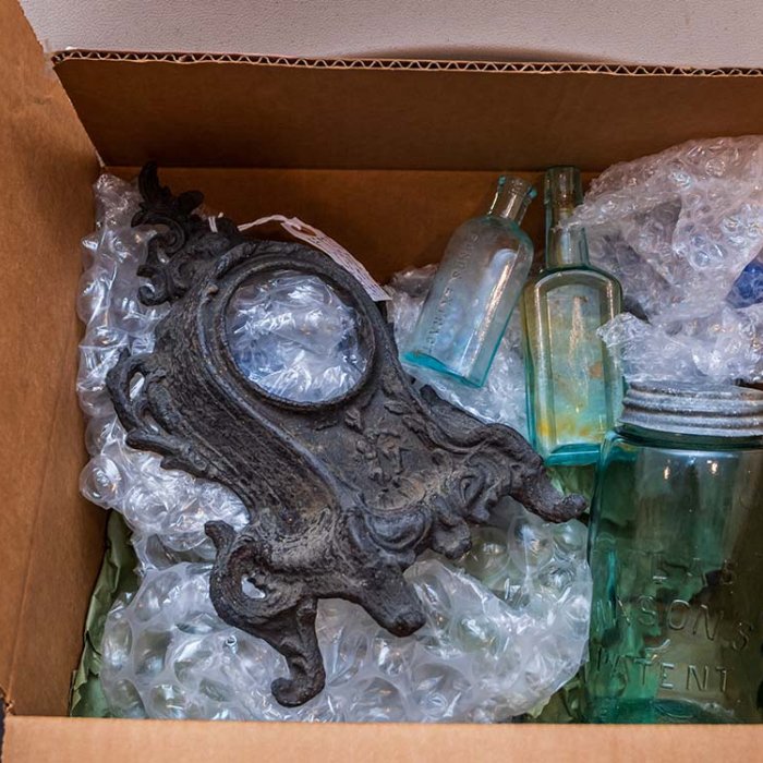 A cardboard box with bubble wrap containing an iron object that looks like a clock body and some antique bottles and glassware.