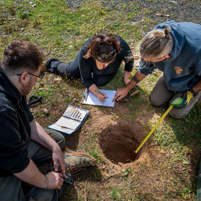 Three people kneel around a hole in the ground while one takes notes.