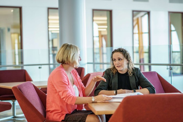 Two people in professional attire are seated at a table, engaging in discussion with documents in front of them.