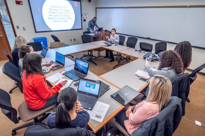 Student in a square shape desk wise at their laptops listening to their professor give a lecture.