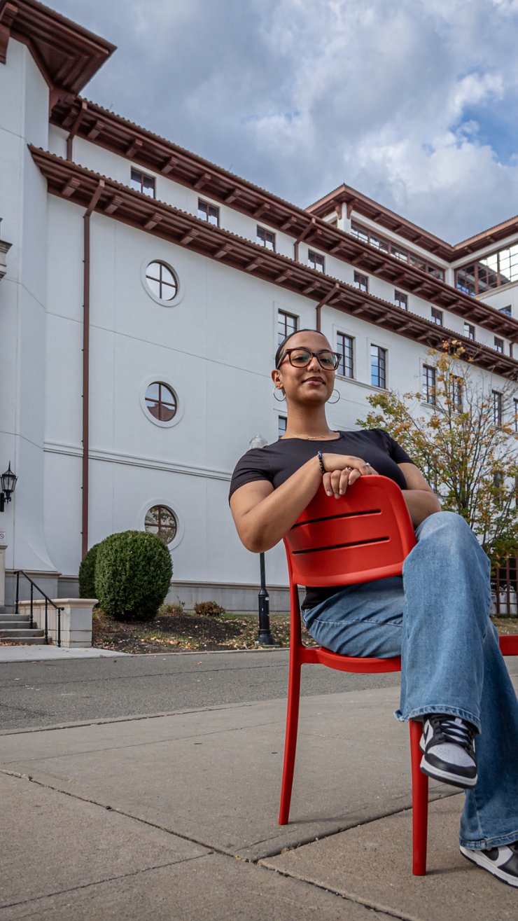 A student sits in a red chair in front of an academic building.