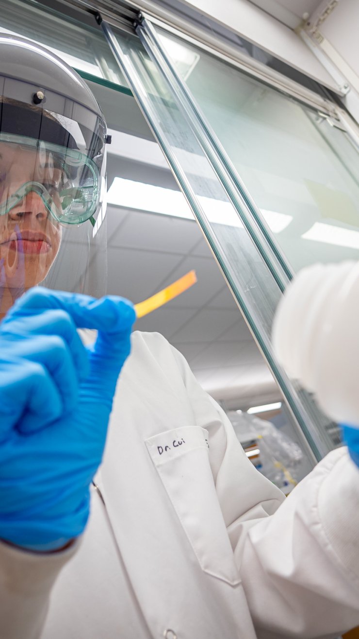 Student in full lab safety gear, including face shield, goggles and gloves, using a test strip while holding a labeled chemical bottle.