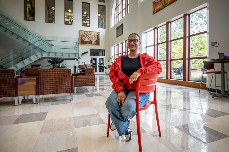 A female student sits on a red chair in the lobby of an academic building. 