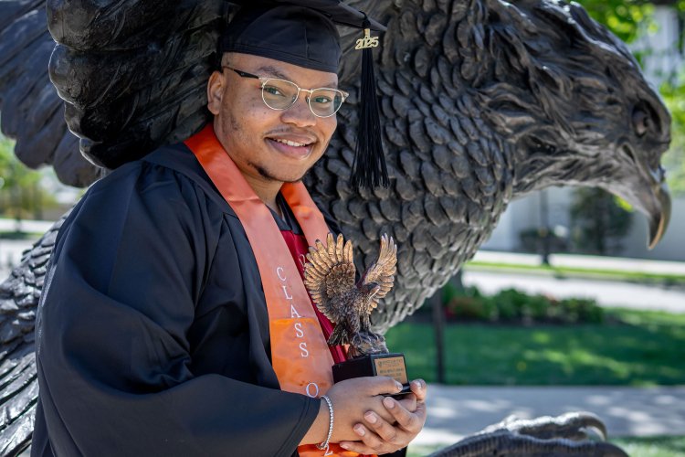 Emmanuel Ozias poses in cap and gown in front of the Red Hawk statute holding a trophy shaped like a red hawk.