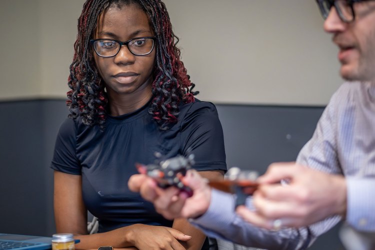 A professor demonstrates a small robot constructed from household items to a student seated nearby.