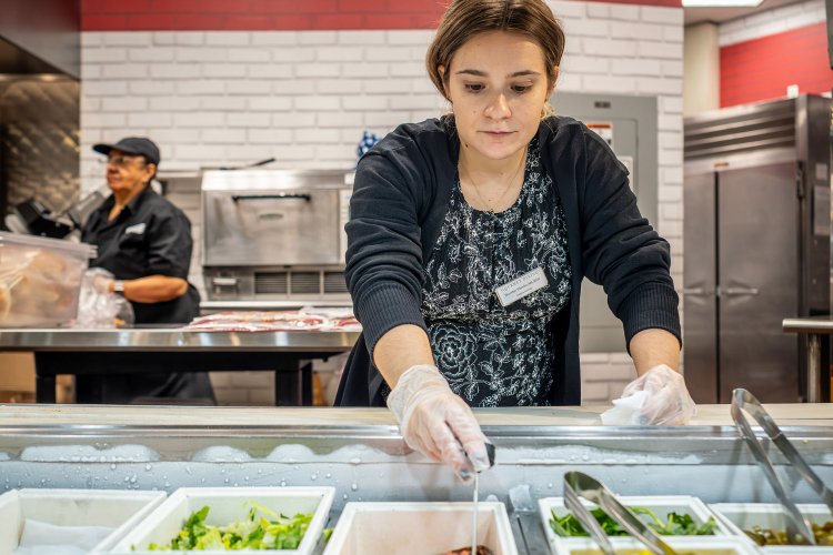 Woman in front of a bar of food with gloves on reaching in.