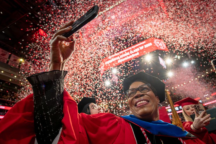 A doctoral graduate in a red gown and black doctoral cap smiles and raises a phone to capture the moment as red and white confetti rains down inside Prudential Center.