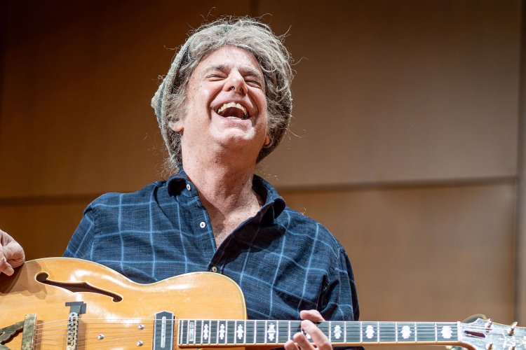 Pat Metheny laughs on stage as he holds a guitar in his lap.