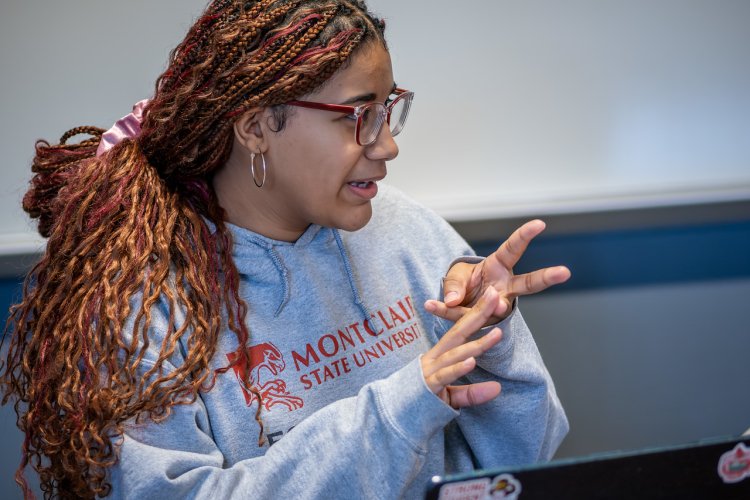A student with braided hair sits in a classroom wearing a Montclair State University hoodie, gesturing with their hands while participating in class.