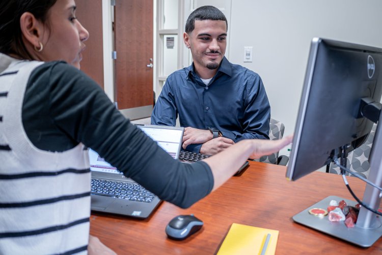 Advisor meets with a student at a desk, reviewing information on a computer screen.