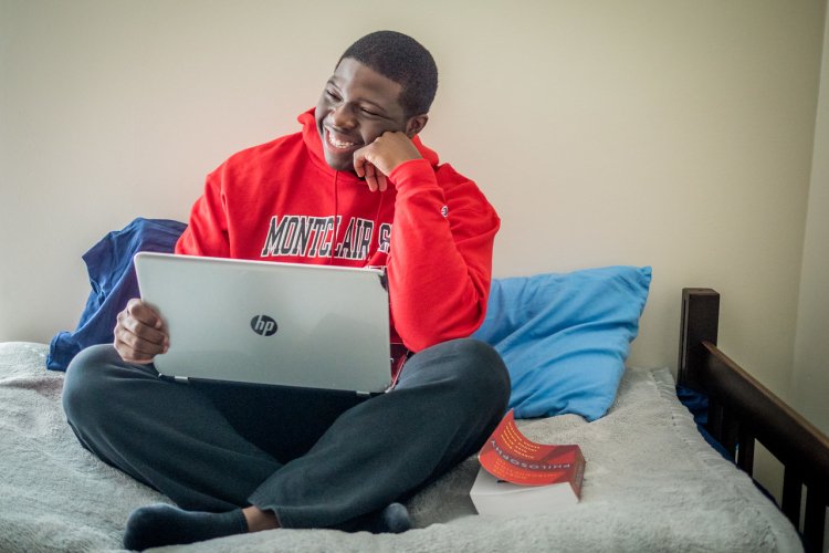 A student wearing a Montclair State University hoodie sits on a bed working on a laptop.