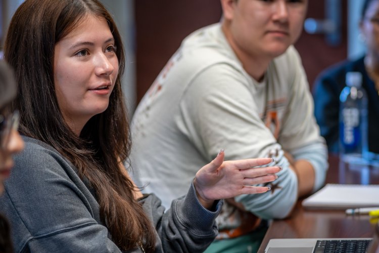 A student with long brown hair in a gray sweater gestures with her hand while speaking during a classroom or group discussion. Another student is seated nearby, focused on the conversation.