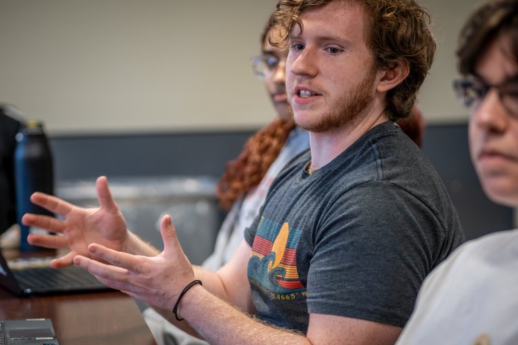 A student in a graphic t-shirt sits at a conference table, gesturing with both hands while speaking during a class or group discussion.