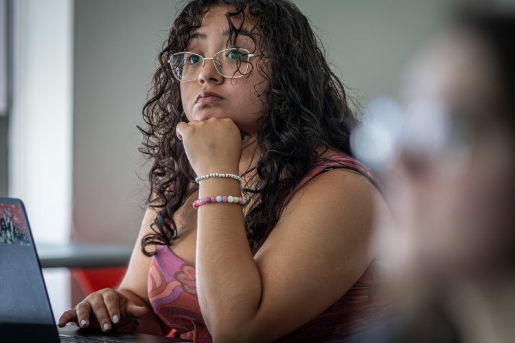 A student working on a laptop in a classroom, resting their chin on their hand and listening attentively during class.