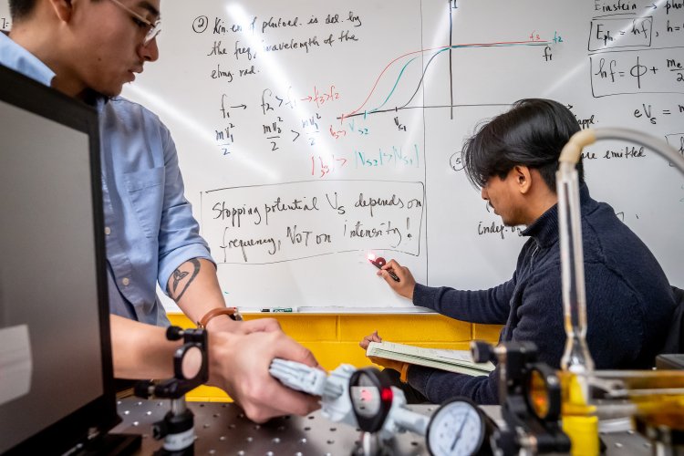 Two students work in a physics lab, with one seated at a whiteboard drawing graphs and writing equations while the other stands beside an optical bench covered with lenses, gauges, and equipment in the foreground.