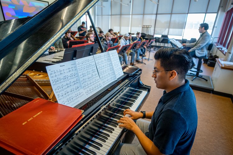 Music student playing a grand piano in a rehearsal room, with sheet music on the stand and an ensemble and conductor in the background.