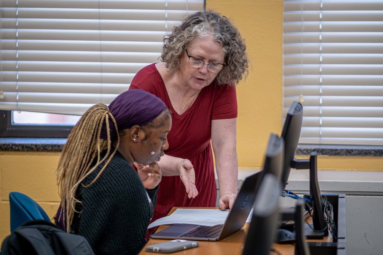 A professor in a computer lab works with a student, referring to the student's notes