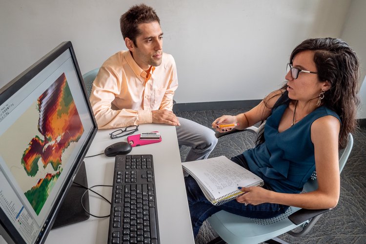 Student and professor sit at a desk discussing notes while a computer monitor displays a colorful scientific data map.