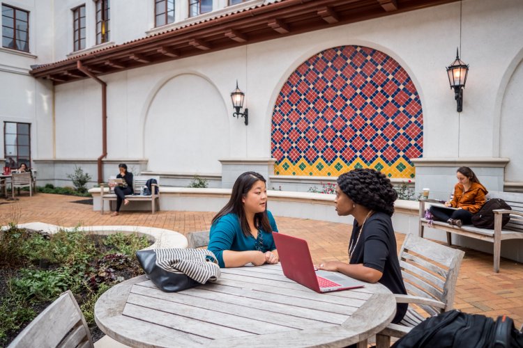 Two women sit talking at an outdoor table in a courtyard.