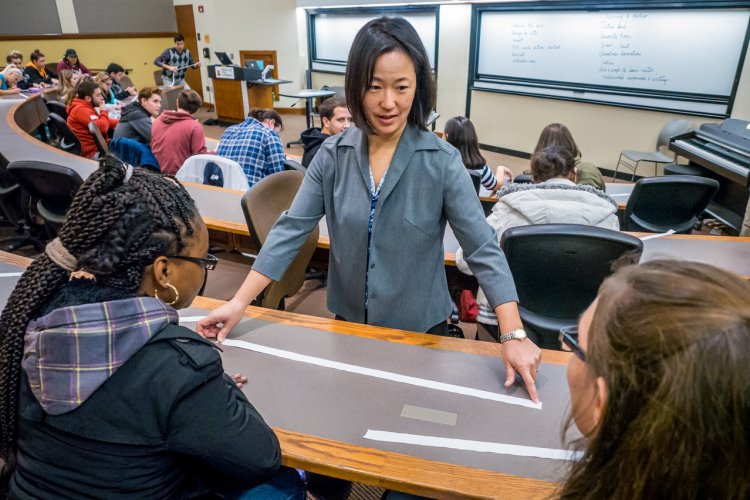 A professor stands at the front of a curved lecture hall aisle, leaning over a long strip of paper taped to a desk during a creative math activity, gesturing as two students seated nearby look on, while other students work at desks in the background.