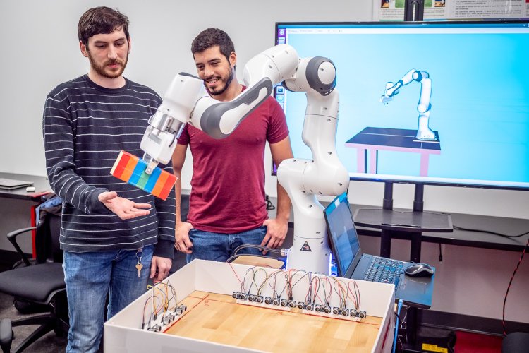 Two students stand beside a white robotic arm that is holding colorful stacked blocks over a sensor-lined wooden testbed, while a large monitor behind them shows a 3D simulation of the same robot.