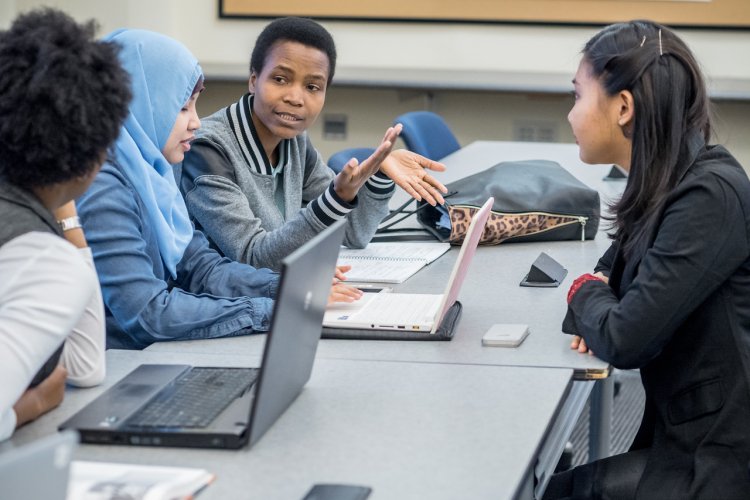 Graduate students at a table discussing over laptops and hand gestures.