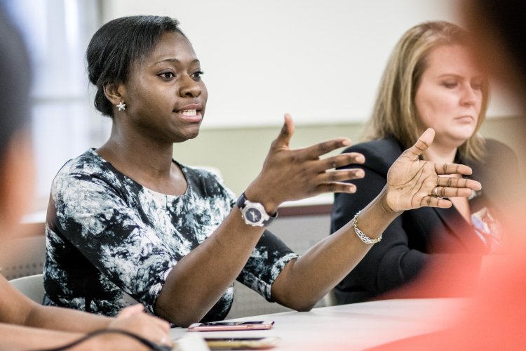 Graduate student speaking and gesturing during a classroom discussion, seated at a table with classmates.
