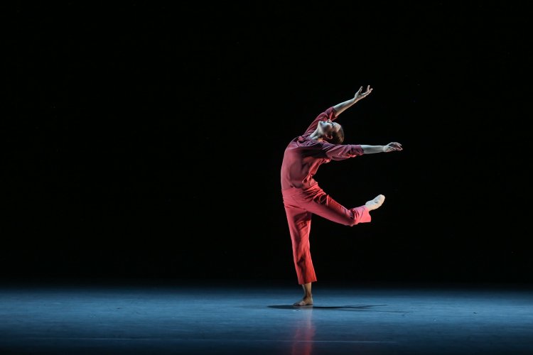 Dancer in a red costume performing a leap with arched back and outstretched arms on a dark stage.