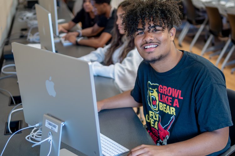 Bloomfield  student smiling at a desktop computer in a campus lab, with classmates working at computers in the background.