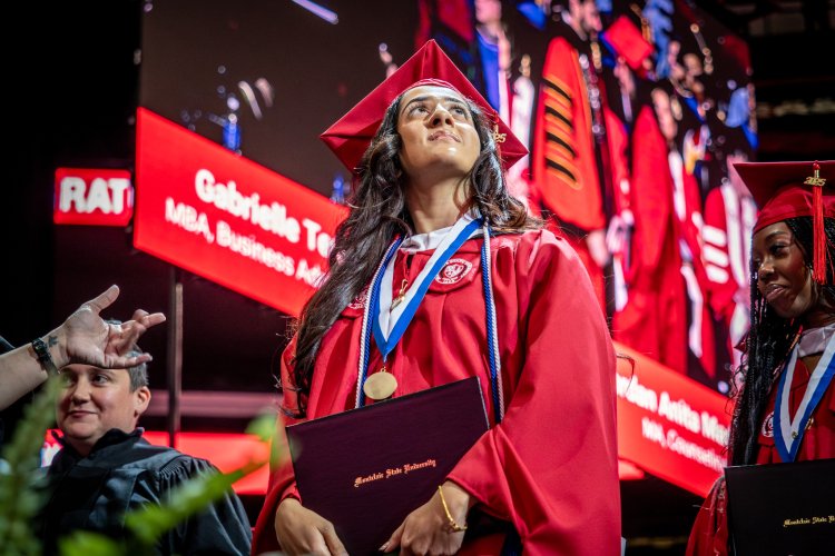 A graduate in a red cap and gown holds a diploma and medal on stage at commencement.