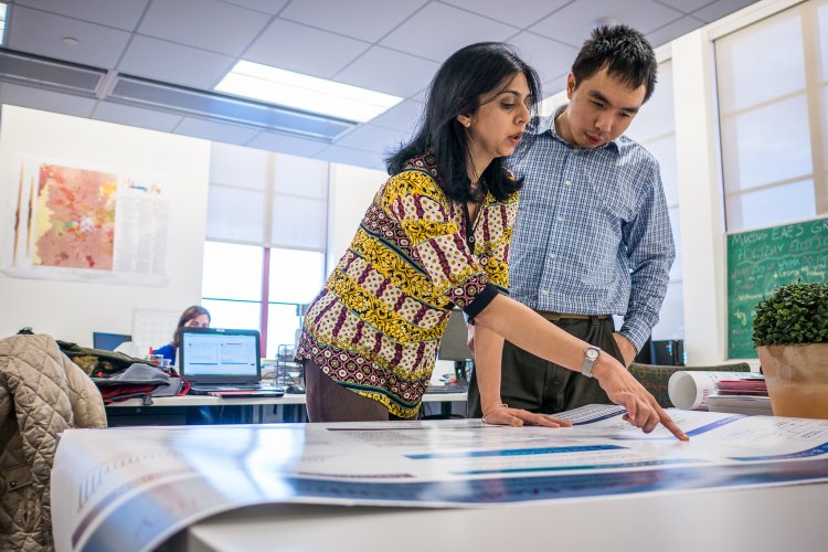 Professor and student stand at a table in an office, reviewing and pointing to large printed charts spread out in front of them.