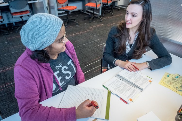Student and professor sat down at a table discussing with pens and papers in front of them.