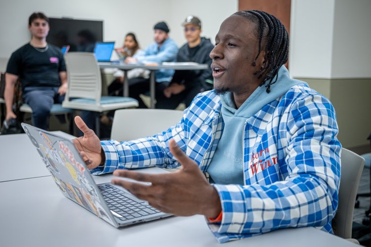 A student in a plaid jacket speaks during a class discussion, gesturing with hands while sitting at a laptop, with classmates working together at a table in the background.