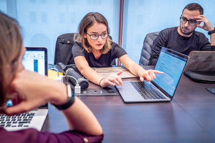 Students sit around a conference table with laptops while one student leans forward and points at a shared laptop screen.